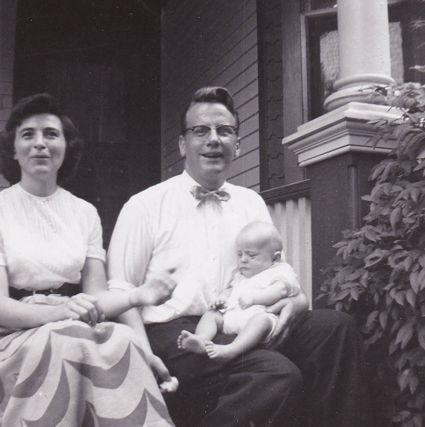Man and woman sitting side by side on a front porch of a home. The father is holding the young baby on his lap.