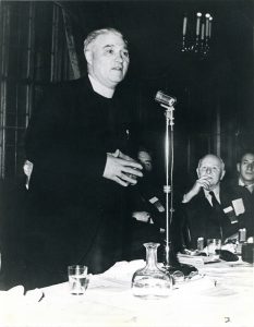 Photograph of Father Moses Coady speaking at a microphone with other men seating in the background