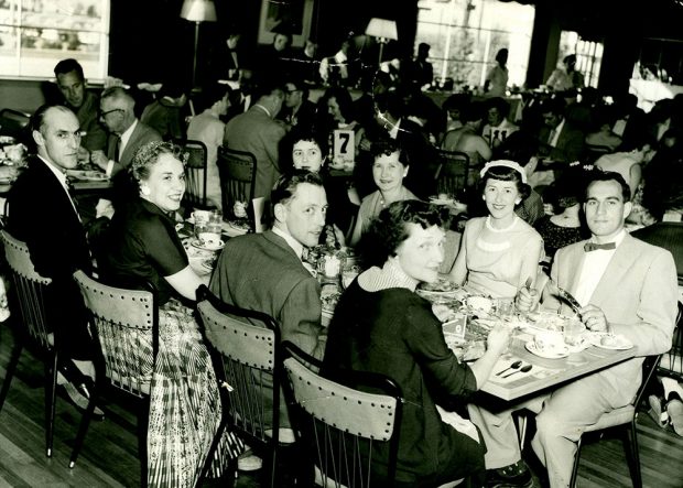 Black and white photograph of a group of men and women sitting at a table eating at a gathering