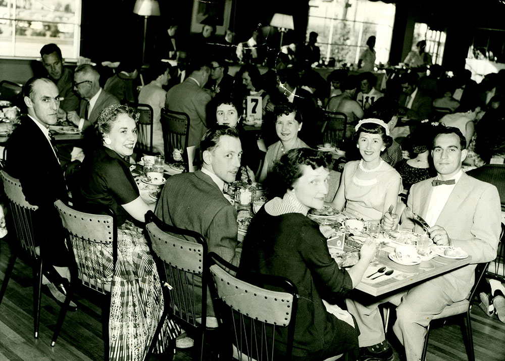 Photo noir et blanc d’un groupe d’hommes et de femmes attablé et mangeant lors d’une réunion.