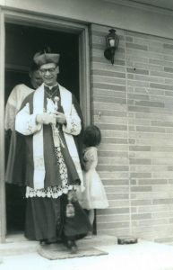 Black and white photograph of a priest walking out of the front door of a house. There is a child standing in the doorway as well and a man walking out behind