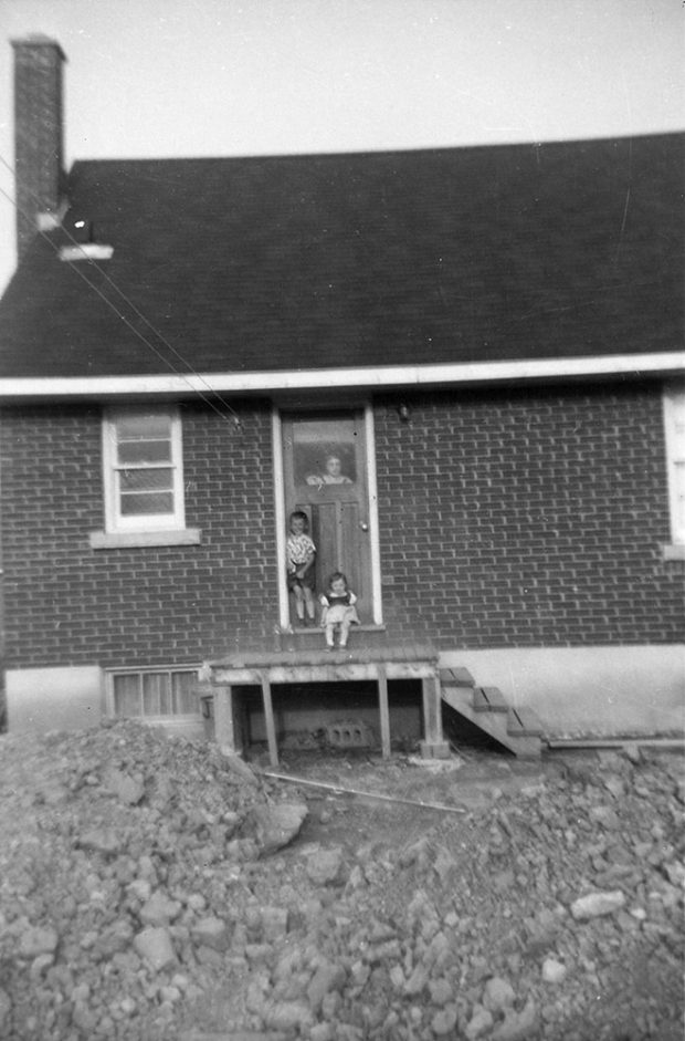 black and white photograph of 2 children out side the back of a house and a woman at the door. House in under construction