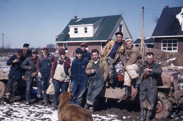 A Colour photograph of a group of men taking a break at a construction site. A dog in the foreground.