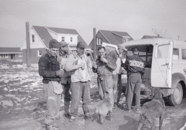 A black and white photograph of a group of men taking a break at a construction site. Two dogs in the foreground.