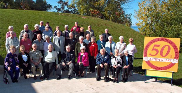 Colour photograph of a group of people standing and sitting in front of a hill and beside a sign that reads Grimsby Homebuilders Housing 50th Anniversary Co-op