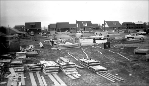 Black and white photograph of a construction site. Piles of materials are laid out around the site