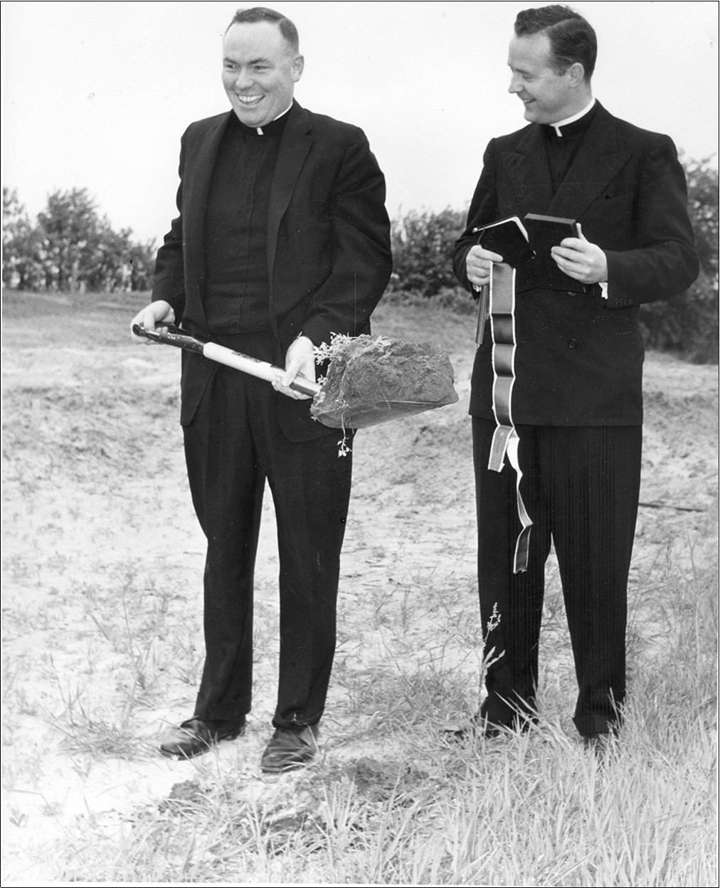 Black and white photograph of two priests at a ground breaking event. One has a shovel the other has a book.