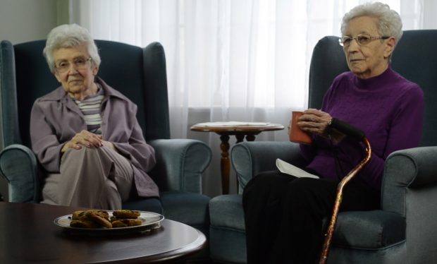 Colour photograph of two women seated in green chairs facing diagonally the one on the right has a cane on her arm and a cup in her hand