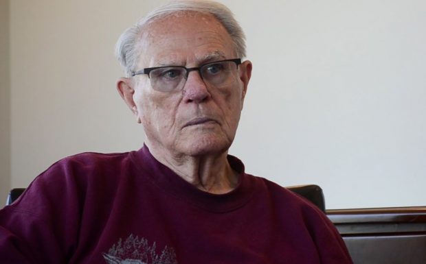 colour photograph of a man in glasses sitting in a chair in a maroon shirt