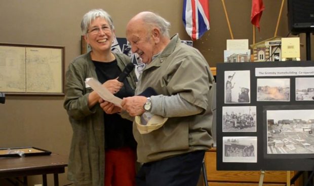 colour photograph of a woman on the left holding a microphone for a man who is reading a paper