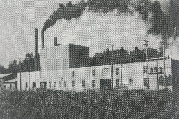 Black and white photo of a long factory building with three chimneys, one of which belches smoke.
