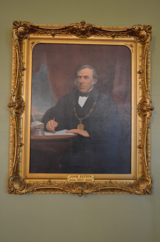 Official portrait painting of Kingston's Mayor John Breden circa 1867, wearing his mayoral robe and chain, seated at his desk holding a pen and paper