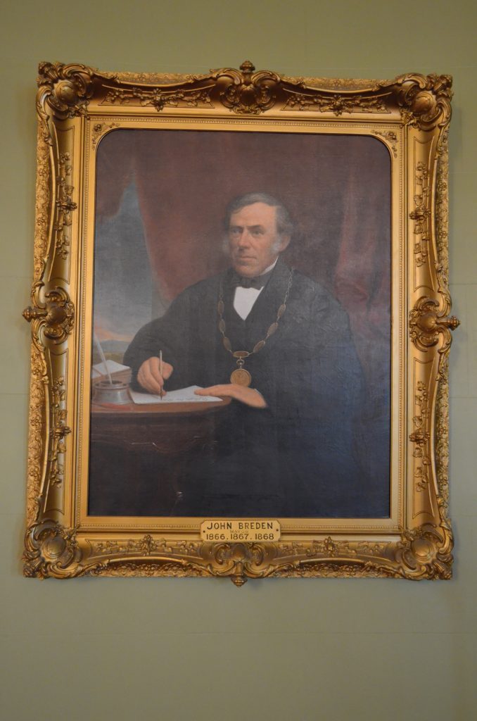 Official portrait painting of Kingston's Mayor John Breden circa 1867, wearing his mayoral robe and chain, seated at his desk holding a pen and paper