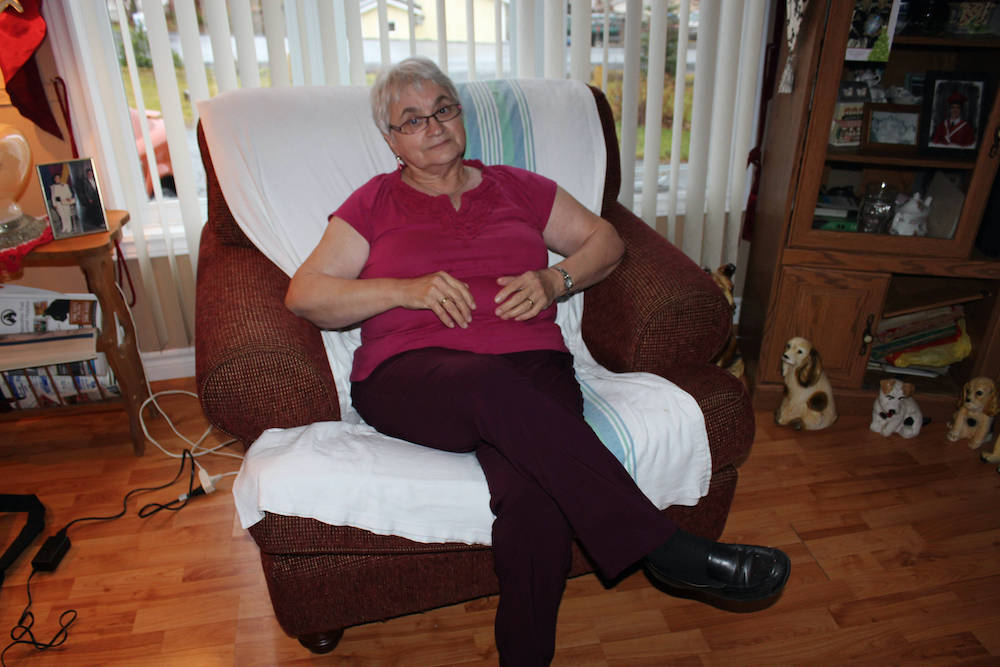 A colour photograph of a woman with grey hair and glasses, taken indoors. The woman is seated in a comfortable easy chair, in front of a window.