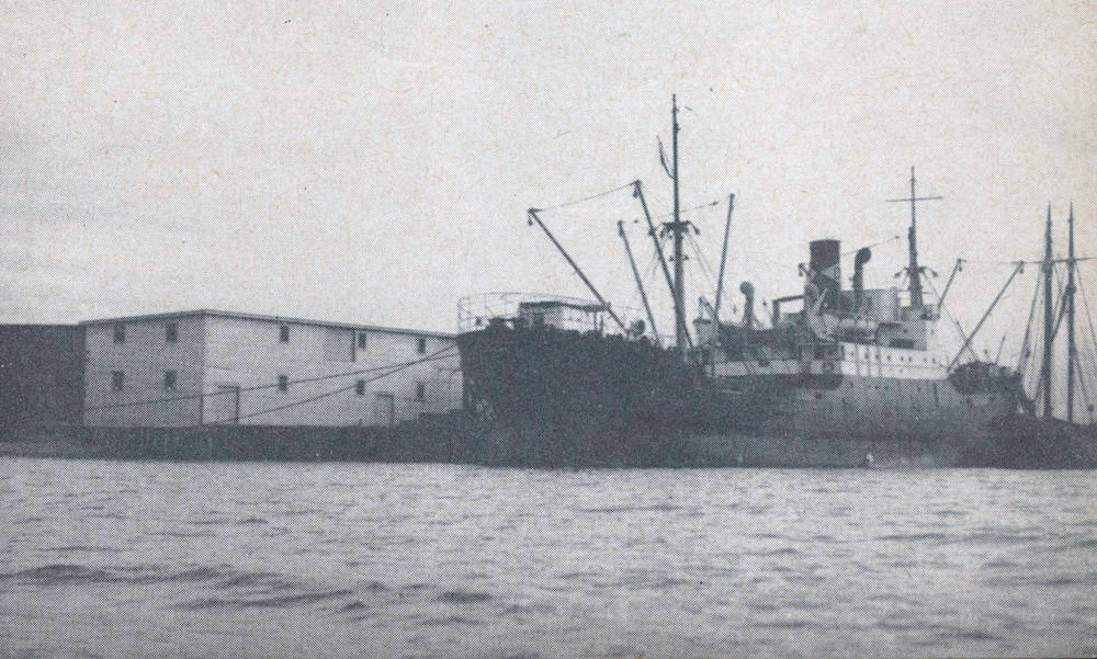 Black and white photograph of a steamship unloading coal, with a white two-storey warehouse in the left background of the photo.