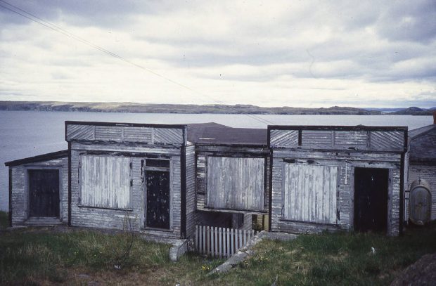Colour photograph of a weathered and dilapidated store, with boarded up windows. The ocean and a distant shoreline are in the background of the photo.
