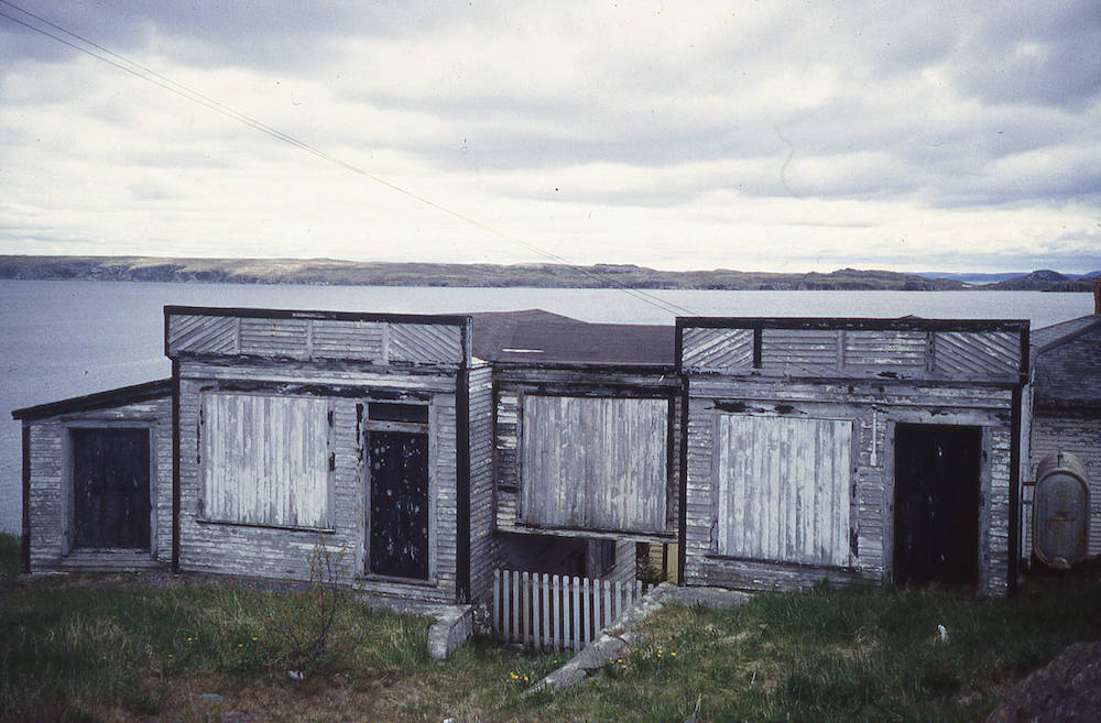 Colour photograph of a weathered and dilapidated store, with boarded up windows. The ocean and a distant shoreline are in the background of the photo.