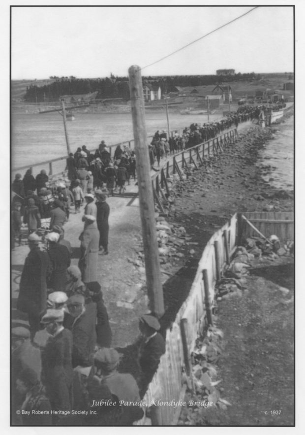 Black and white archival photograph of a causeway and bridge over a body of water, with a crowd of people marching in a parade away from the camera.