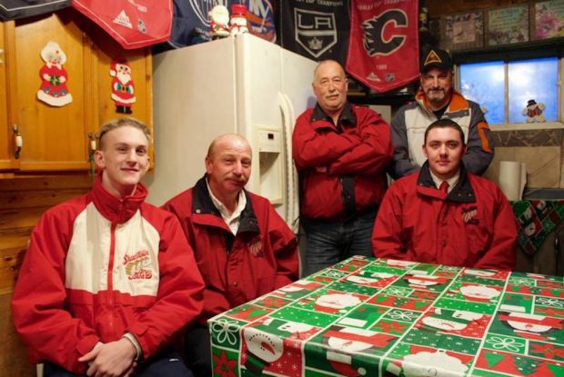 A colour photograph of three men are seated at the kitchen table and two men standing behind them.