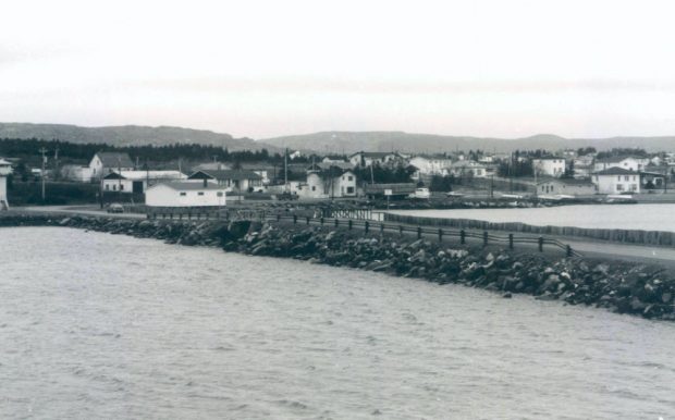 Black and white archival photograph of a causeway and bridge over a body of water, with the shoreline and houses beyond, and hills in the distant background.