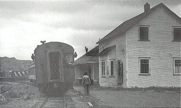 Black and white archival photograph showing a traincar on railway tracks to the left, with a single storey wooden freight shed and two storey wooden train depot to the right.