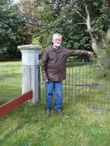 A colour photograph of a man with white hair and white beard, dressed in a leather coat and jeans, standing in front of an iron gate, with a concrete pillar to the left of the photo.