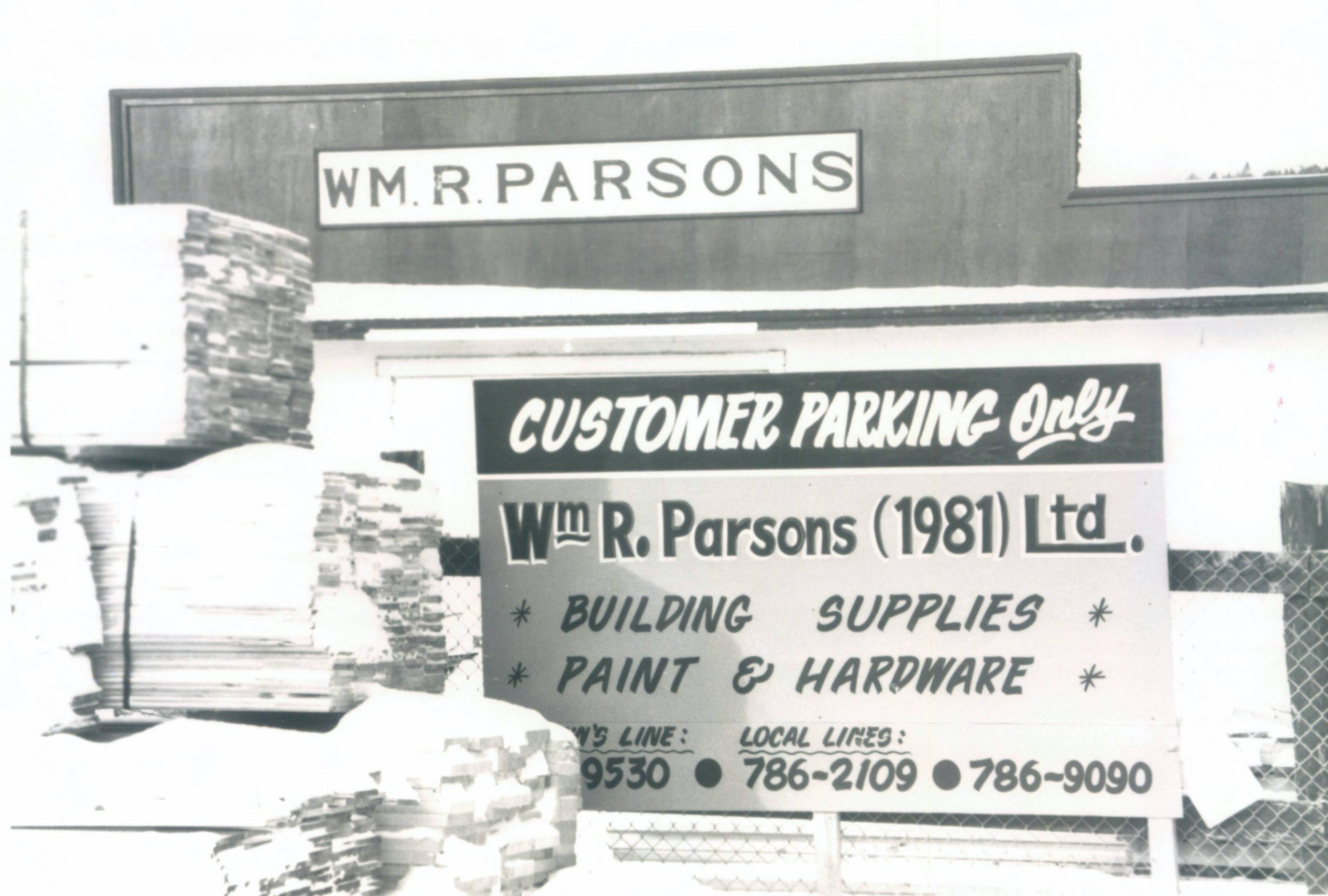 Photographie montrant l’extérieur d’un site commercial avec deux grands panneaux peints affichant le nom de l’entreprise, William R. Parsons. 