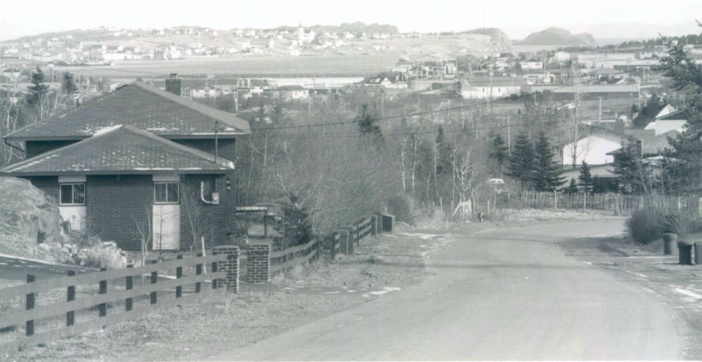 A black and white photograph looking down a road lined with fences. There is a house to the left of the photo, and in the distance, more of the community can be seen, with hills and water behind.