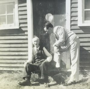 A black and white photo of two men on the front step of Littlejohn's forge. William H. Littlejohn is seated on the step and his grandson, Bill Littlejohn, stands beside him with his hands resting on one knee.