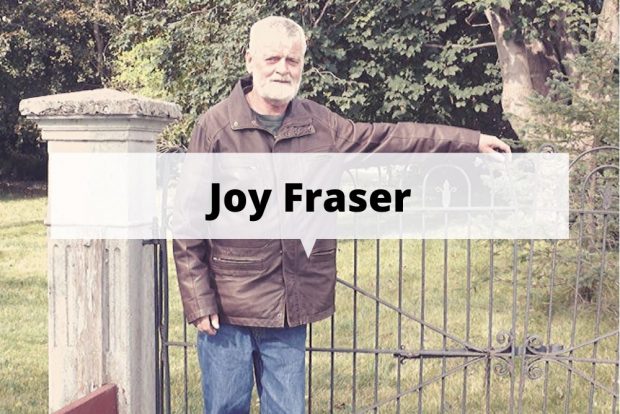 A colour photograph of a man with white hair and white beard, dressed in a leather coat and jeans, standing in front of an iron gate, with a concrete pillar to the left of the photo.