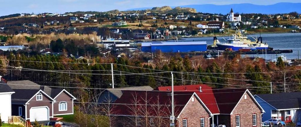 Colour photograph showing a ship in a bay with trees and homes wrapping around the bay in the foreground and background.