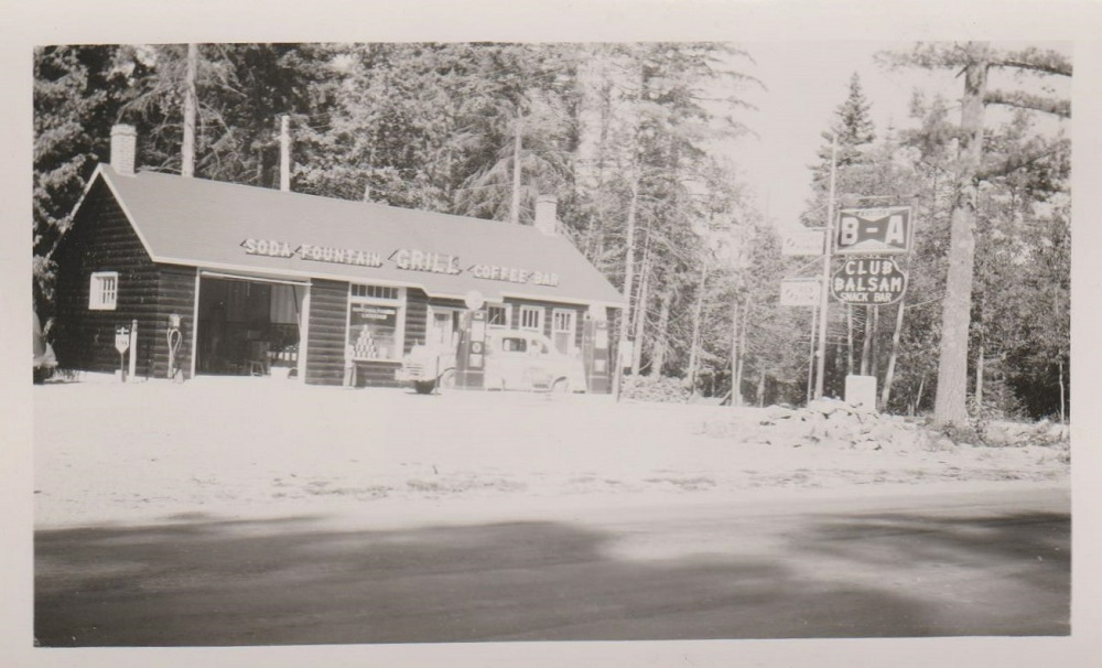 Black-and-white image of log building with gas pumps and vintage car in front; sign at right; and trees in background