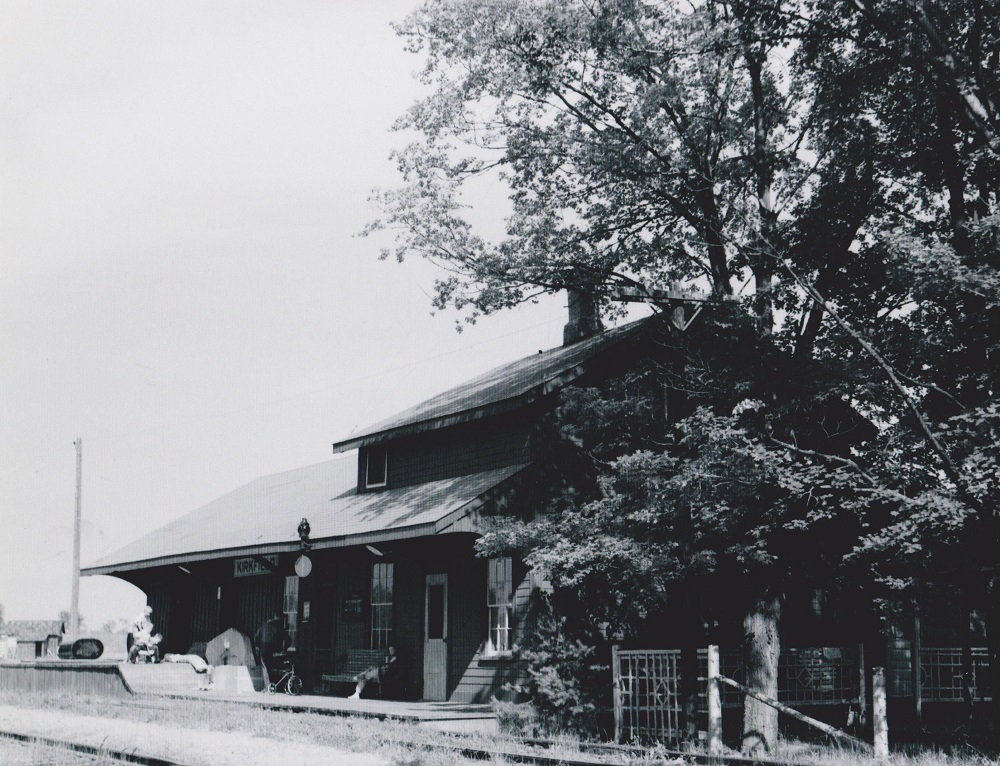 Black-and-white image of railway station with people on platform and large tree growing beside building