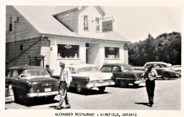 Black-and-white image of building with people and vintage cars in foreground and trees in background
