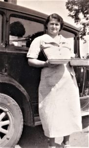 Black-and-white image of woman in white dress carrying two pies in front of vintage car