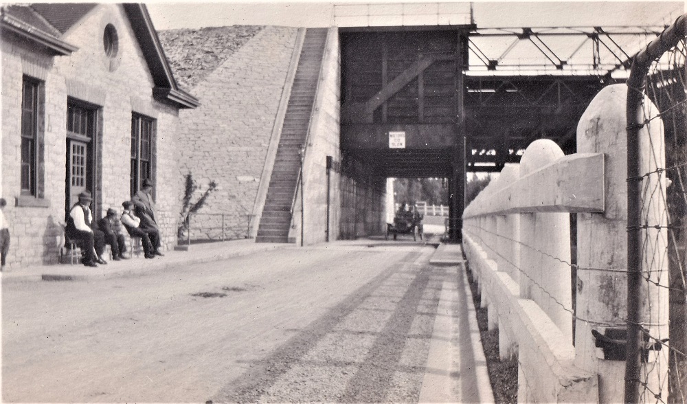 Black-and-white image of vintage car passing through tunnel beneath lift lock with buildings and people in foreground