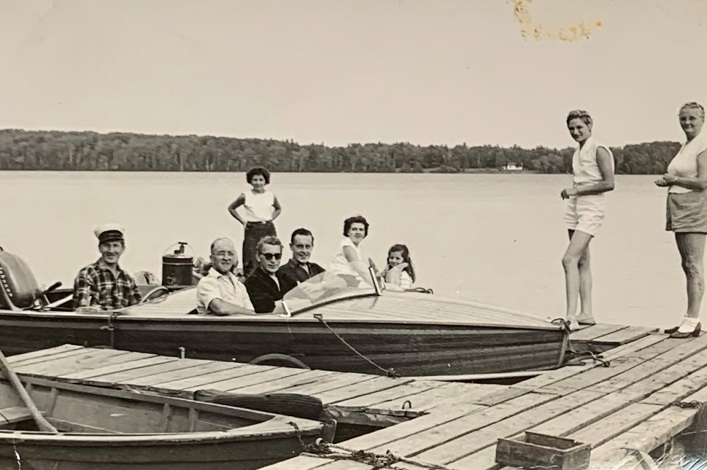 Black-and-white image of people in wooden motorboat at dock with lake in background
