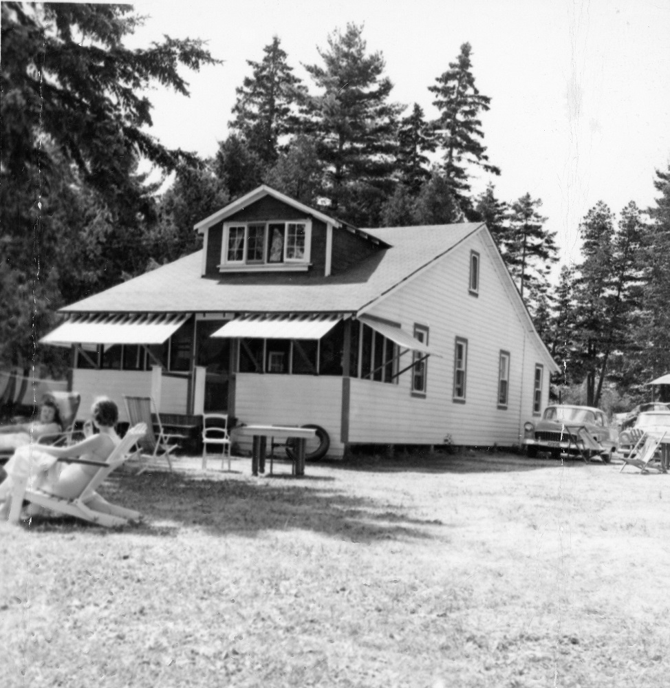 Black-and-white image of white frame building with people on lawn furniture in foreground and vintage car in background