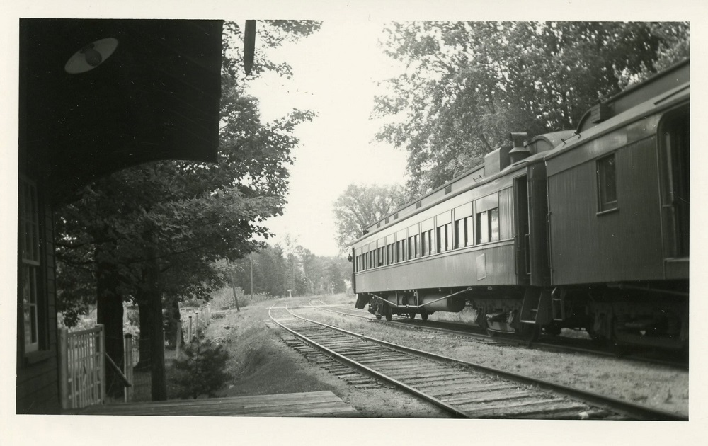 Black-and-white image of train with tracks in foreground and trees in background