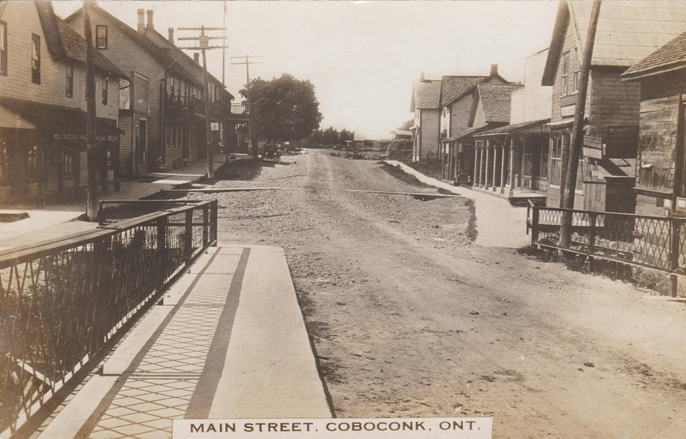 Black-and-white image of a dirt road with bridge in foreground and buildings on either side of road