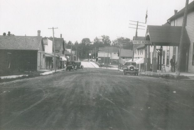 Black-and-white image of streetscape with vintage cars parked in front of buildings and hydro poles lining either side of street