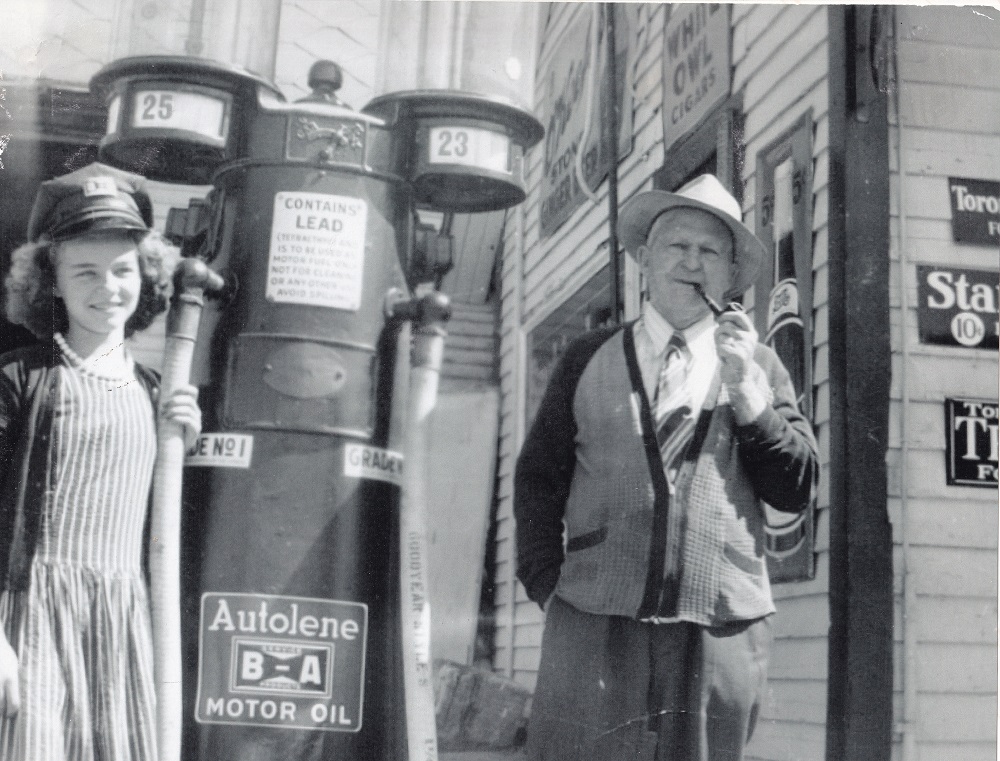 Black-and-white image of young woman in uniform and older man standing in front of vintage gasoline pump