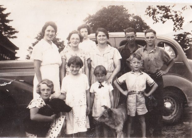 Black-and-white image of adults, children, and two dogs in front of vintage car with trees in background