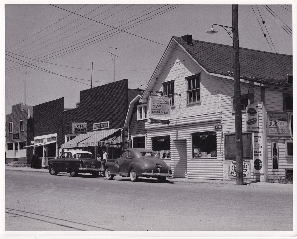 Black-and-white image of buildings decorated in signs, with two vintage cars parked in foreground and electrical wires above