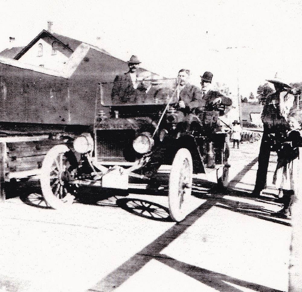 Black-and-white image of people in vintage car crossing a bridge with building in background