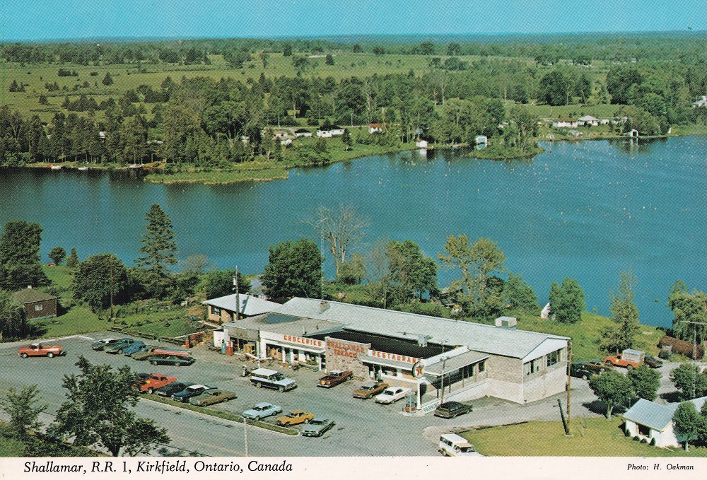 Colour image taken from air showing lake, building, and parking lot with vintage vehicles
