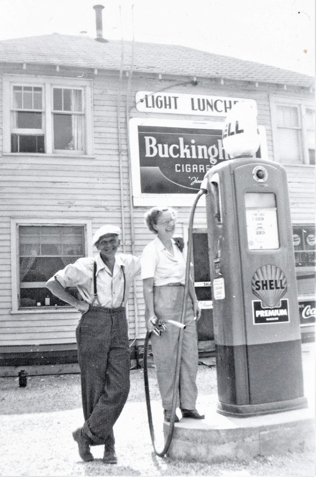 Black-and-white image of two people in front of a gas pump with large wooden building in background