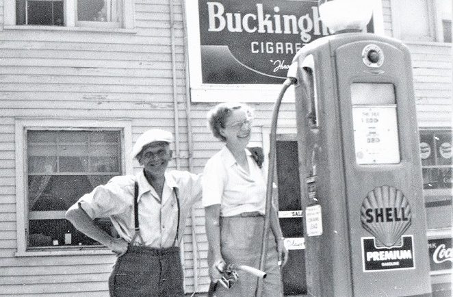 Black-and-white image of two people in front of a gas pump with large wooden building in background