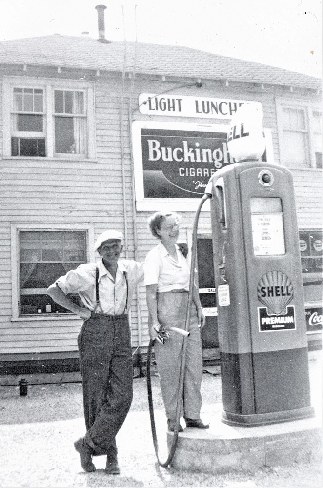 Black-and-white image of two people in front of a gas pump with large wooden building in background