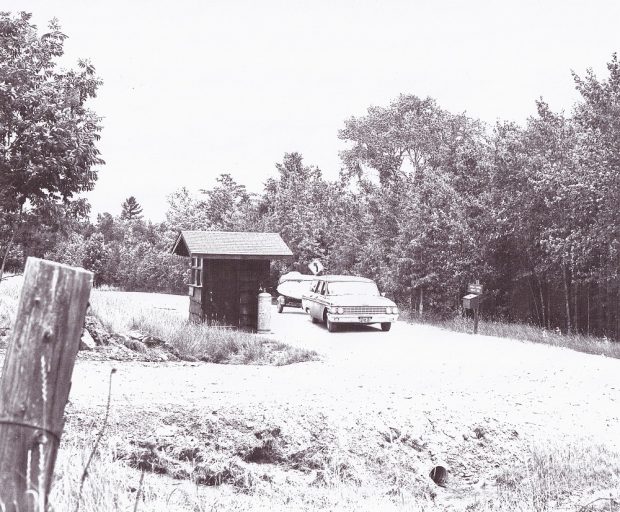 Black-and-white image of vintage vehicle towing boat trailer on dirt road past shack with trees in background